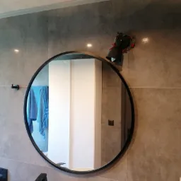 Szare płytki w łazience z okrągłym lustrem, w którym odbija się biała szafka i błękitny ręcznik, a na ścianie wisi czarna lampa z dekoracją kwiatową.