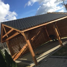 Mik-Mont - Konstrukcja drewniana z czarnym dachem z dachówki bitumicznej w trakcie budowy, widoczny pracownik wewnątrz, drabina oparta o bok, kostka brukowa wokół.