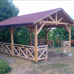 Mik-Mont - Nowa, drewniana konstrukcja z czerwonym dachem w ogrodzie, z drewnianymi barierkami w kształcie krzyży. W tle zielona roślinność.