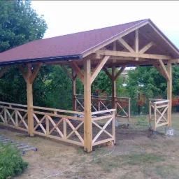 Nowa, drewniana konstrukcja z czerwonym dachem w ogrodzie, z drewnianymi barierkami w kształcie krzyży. W tle zielona roślinność.