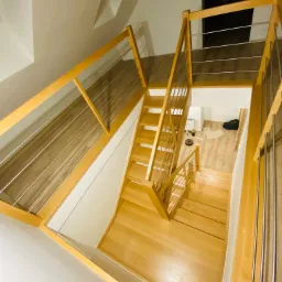 Widok z góry na jasne, drewniane schody z metalowymi poręczami, prowadzące na wyższą kondygnację; widoczny fragment pomieszczenia z robotem sprzątającym.