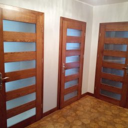 Mik-Mont - Trzy drewniane drzwi wewnętrzne z przeszkleniami, widoczne na tle parkietu ułożonego we wzór kostki, jeden z grzejników jest widoczny po lewej stronie.