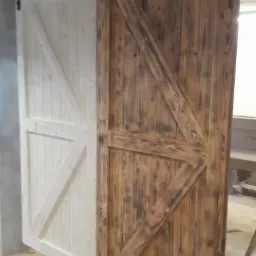 Dwie przesuwne drzwi w stylu rustykalnym z drewna, jedna biała, druga opalana, zawieszone na czarnym metalowym systemie przesuwnym.