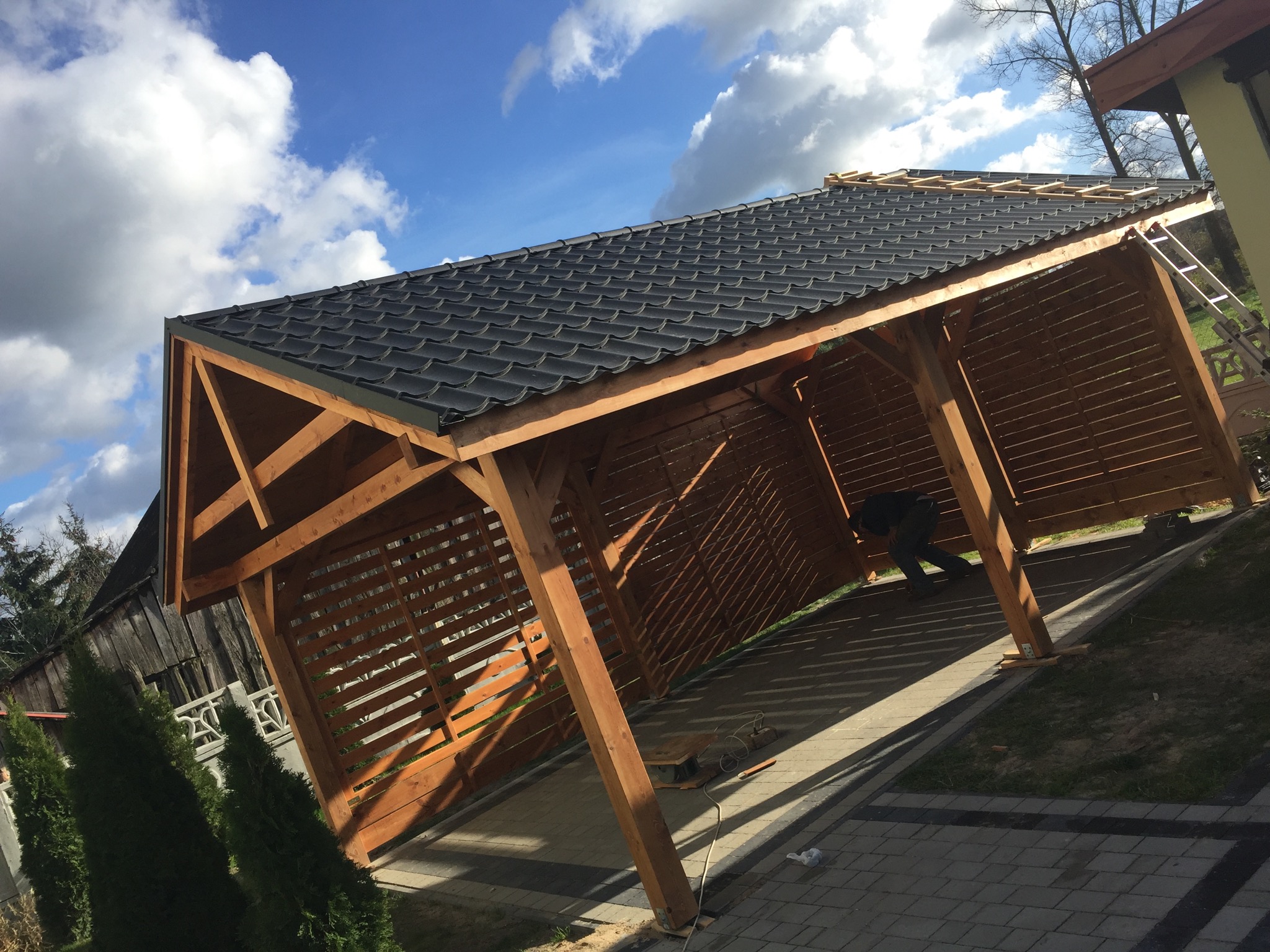 Konstrukcja drewniana z czarnym dachem z dachówki bitumicznej w trakcie budowy, widoczny pracownik wewnątrz, drabina oparta o bok, kostka brukowa wokół.