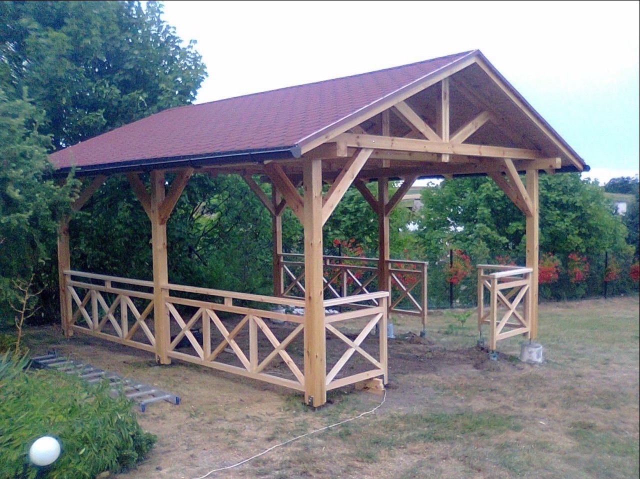 Nowa, drewniana konstrukcja z czerwonym dachem w ogrodzie, z drewnianymi barierkami w kształcie krzyży. W tle zielona roślinność.