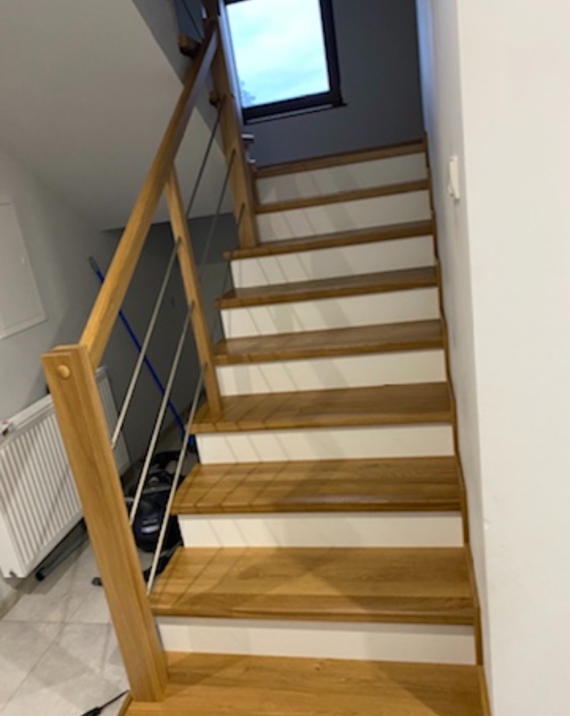 Jasne drewniane schody z białymi podstopnicami i metalowymi poręczami prowadzące do okna dachowego, widok z dołu, ślady drobnych prac wykończeniowych widoczne w tle.