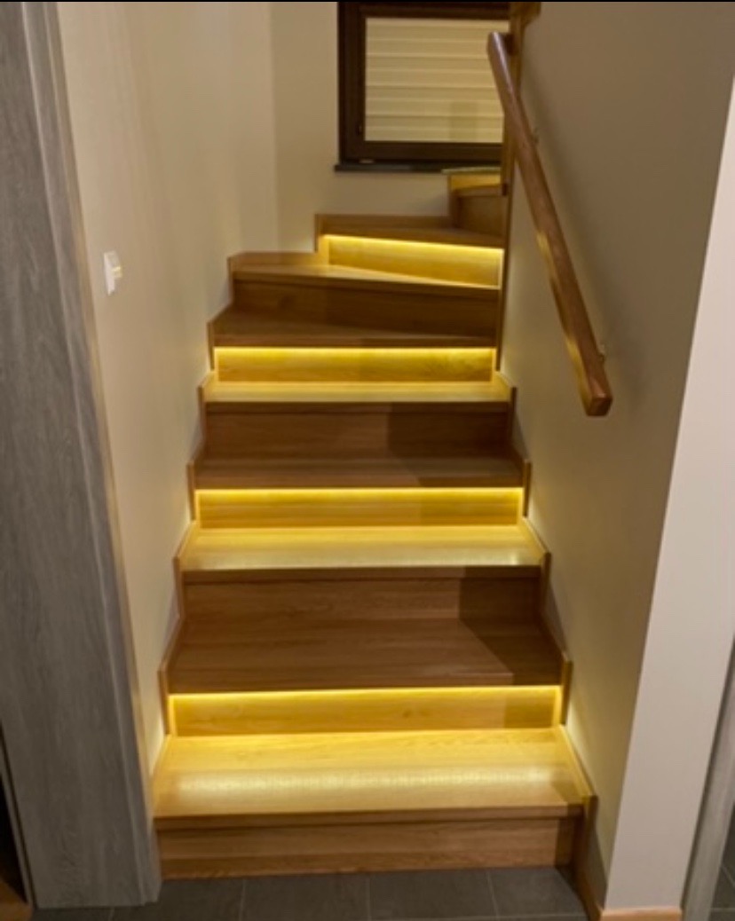 Drewniane schody z podświetleniem LED na każdym stopniu, prowadzące do góry, z drewnianą poręczą po prawej stronie i oknem w tle.