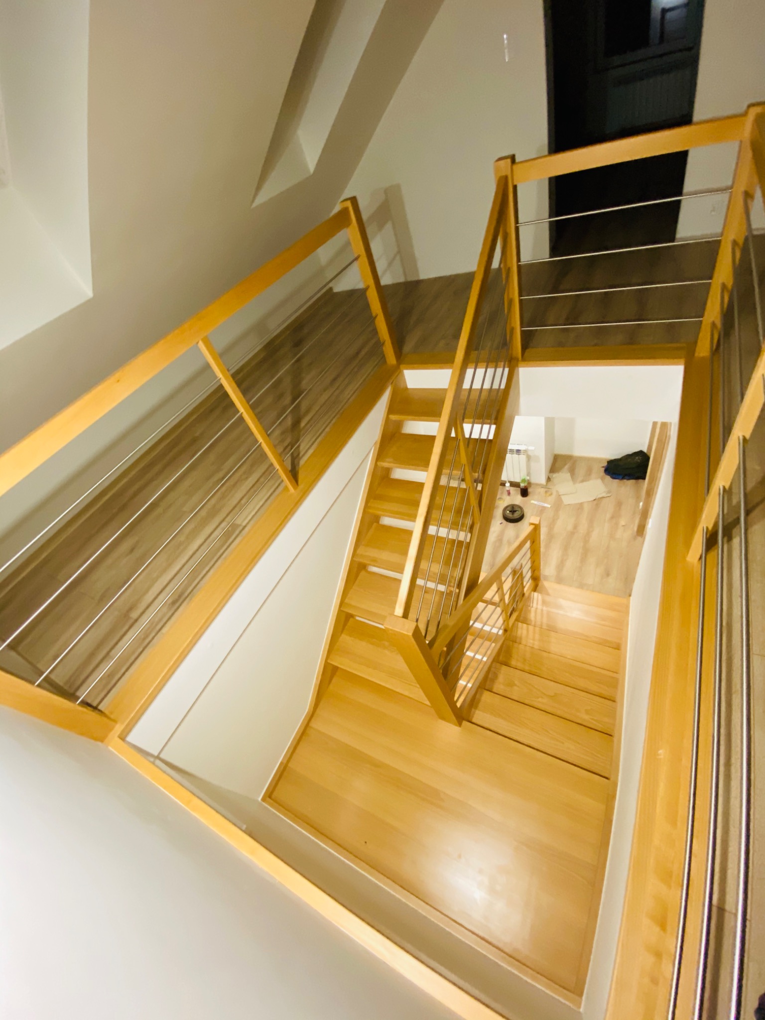 Widok z góry na jasne, drewniane schody z metalowymi poręczami, prowadzące na wyższą kondygnację; widoczny fragment pomieszczenia z robotem sprzątającym.