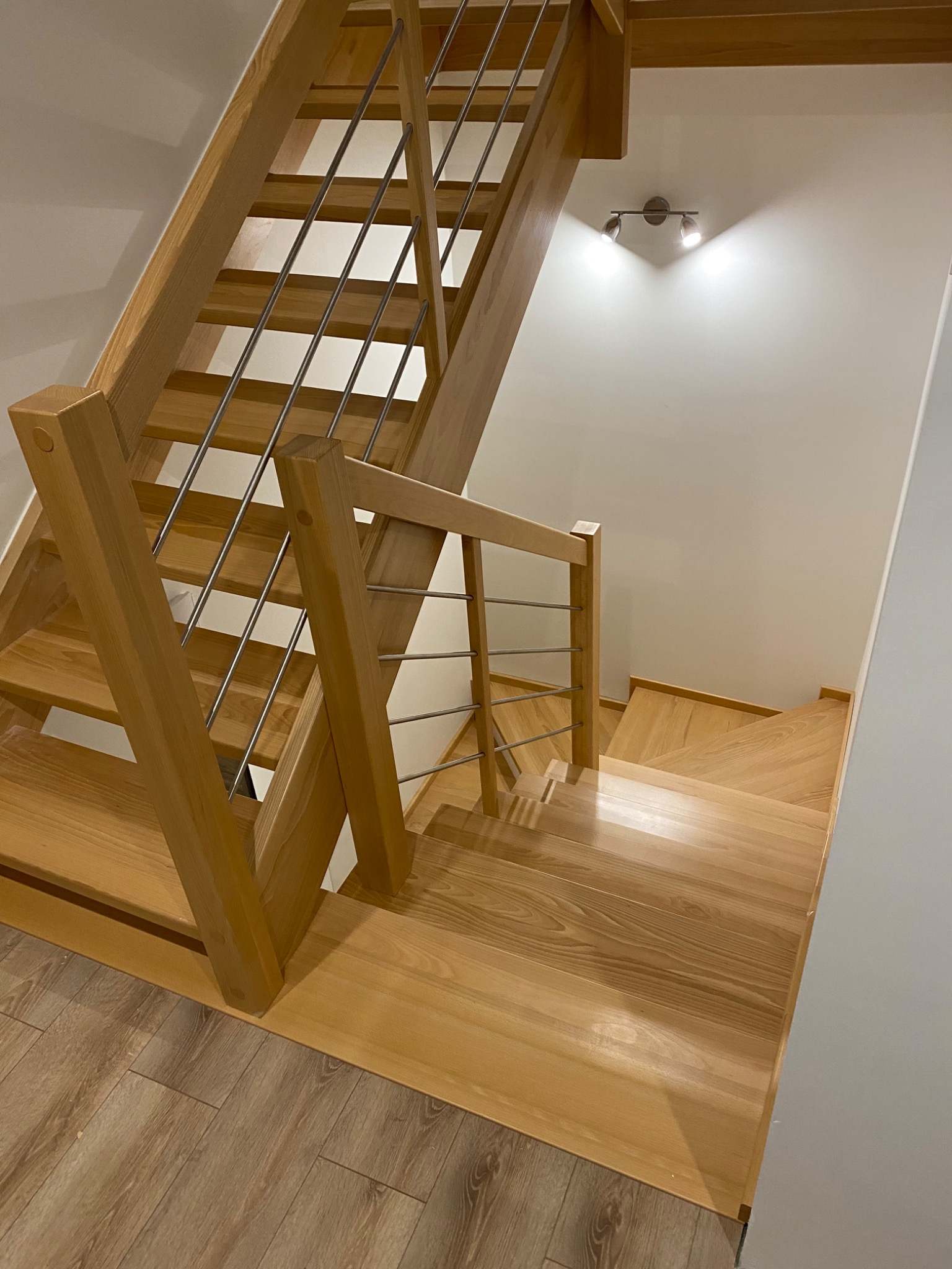 Jasne, drewniane schody z metalowymi poręczami w nowoczesnym wnętrzu, oświetlone lampą punktową na białej ścianie.