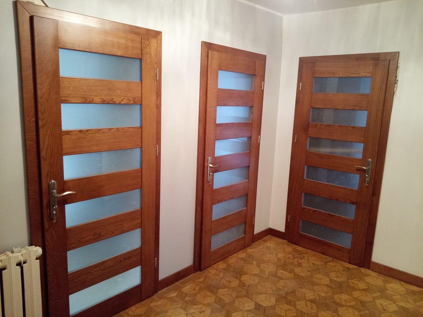 Trzy drewniane drzwi wewnętrzne z przeszkleniami, widoczne na tle parkietu ułożonego we wzór kostki, jeden z grzejników jest widoczny po lewej stronie.
