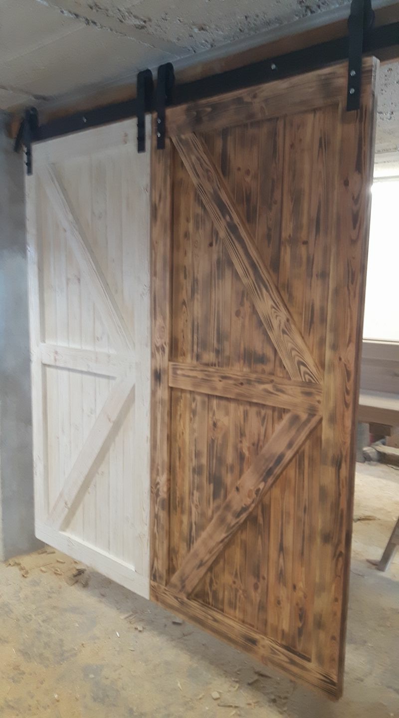 Dwie przesuwne drzwi w stylu rustykalnym z drewna, jedna biała, druga opalana, zawieszone na czarnym metalowym systemie przesuwnym.