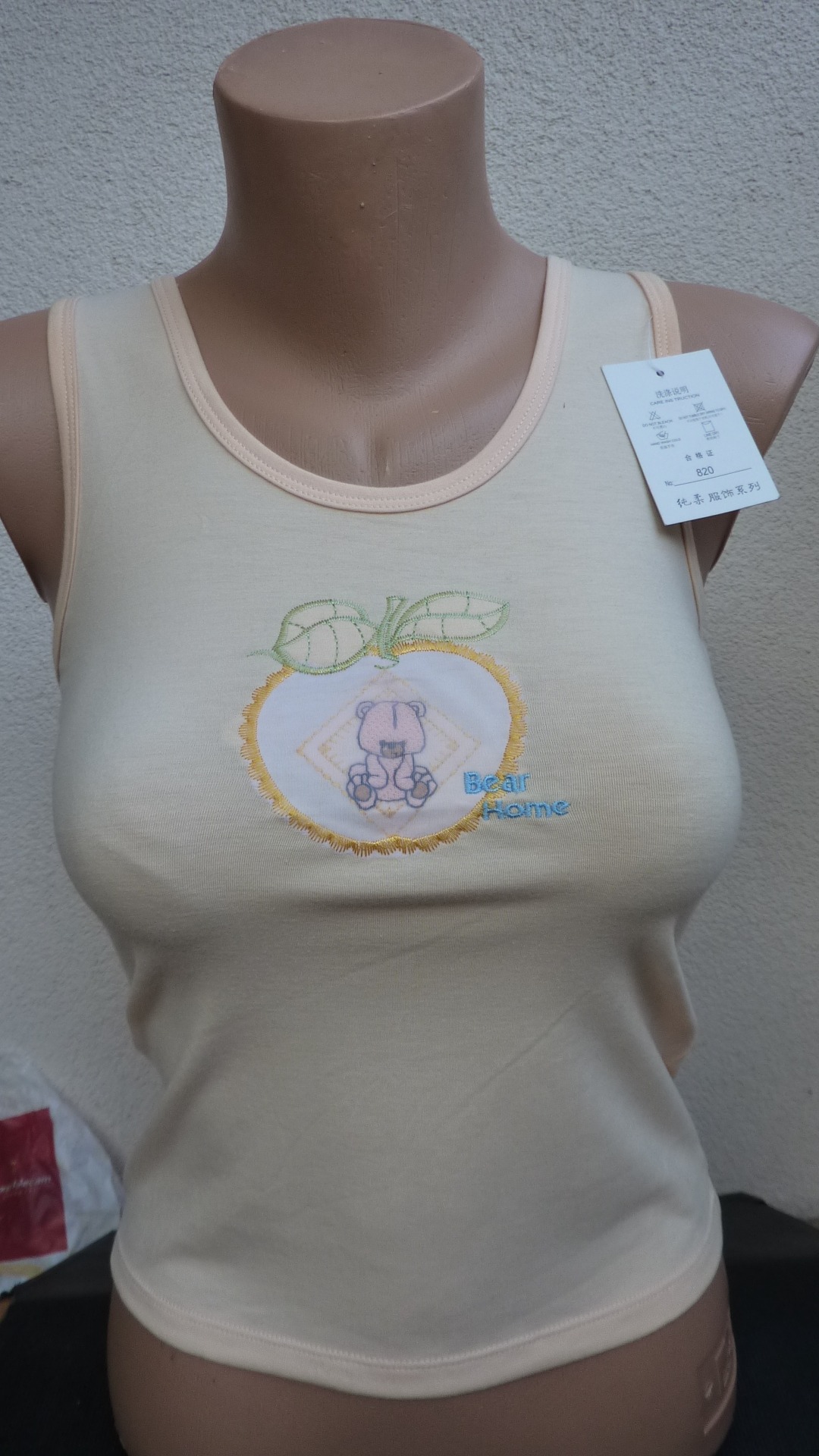 Jasna koszulka na manekinie z aplikacją misia w jabłku i napisem 'Bear Home', widoczna metka z chińskimi znakami.