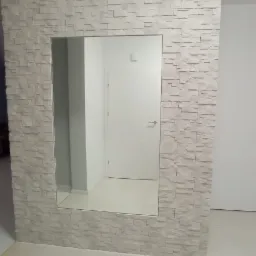 Ściana wykończona dekoracyjną, jasną okładziną imitującą kamień, z wkomponowanym lustrem w przedpokoju. Widoczna biała podłoga z płytek oraz odbicie białych drzwi w lustrze.