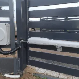 Szara brama wjazdowa z zamontowanym siłownikiem automatycznym marki Nice i skrzynką sterowniczą, widoczny przewód ochronny w ziemi.