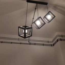 SulanowskiSystem - Czarny, nowoczesny żyrandol z trzema kwadratowymi kloszami zawieszony na suficie z widocznymi rurkami instalacyjnymi.