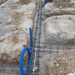 SulanowskiSystem - Wykonane wykopy pod fundamenty z ułożonym zbrojeniem i niebieską rurą karbowaną, widoczny pracownik w niebieskim kombinezonie i czapce.