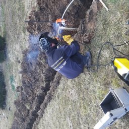 SulanowskiSystem - Spawanie rur w wykopie przez spawacza w masce ochronnej i rękawicach, widoczny sprzęt spawalniczy, żółty przedłużacz i otwarta aluminiowa walizka na trawie.