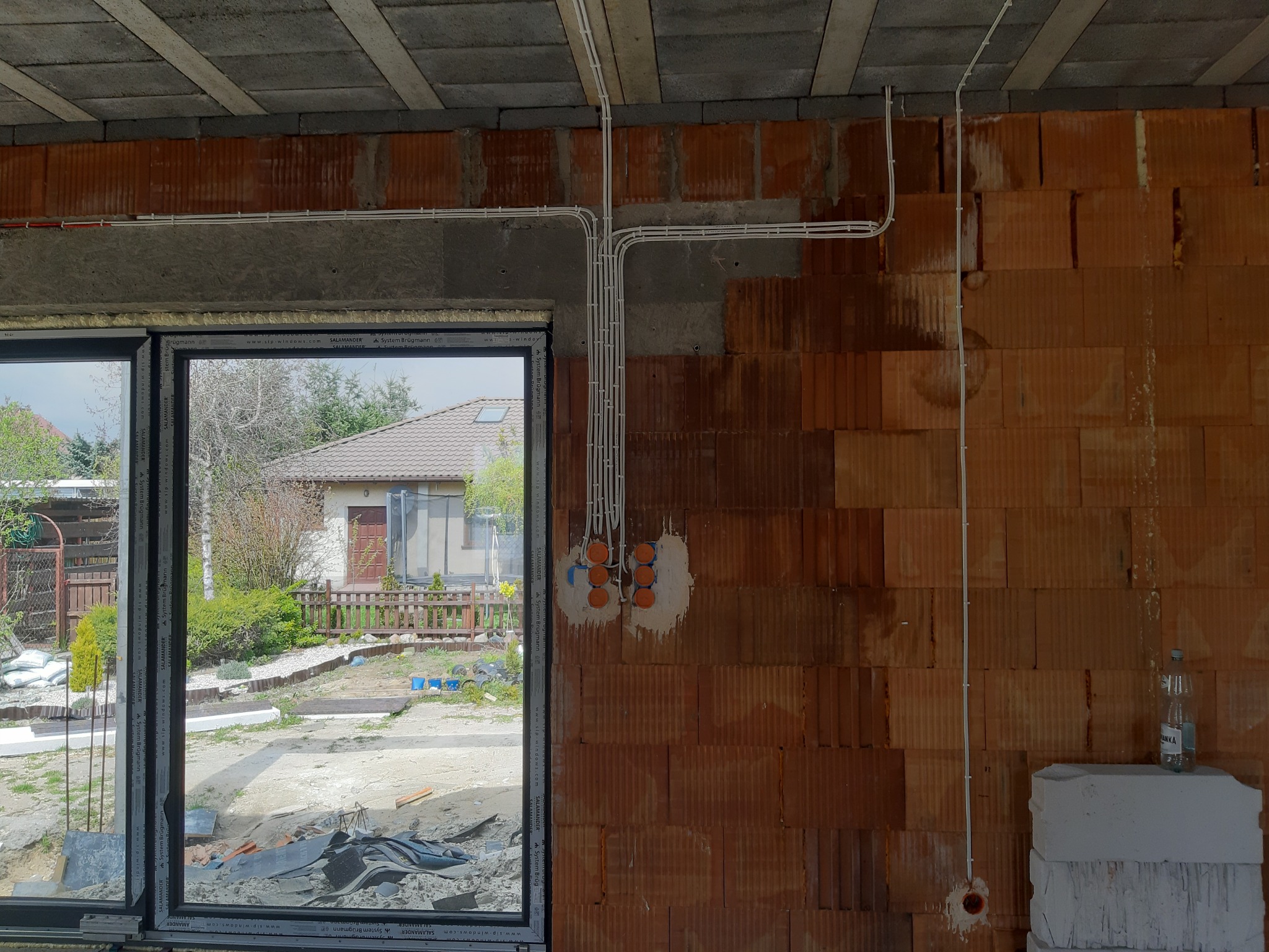 Wewnętrzne okablowanie elektryczne w surowym budynku z widocznymi puszkami instalacyjnymi, przewody prowadzone po ścianie z cegły dziurawki, widok przez okno na dom i ogród.