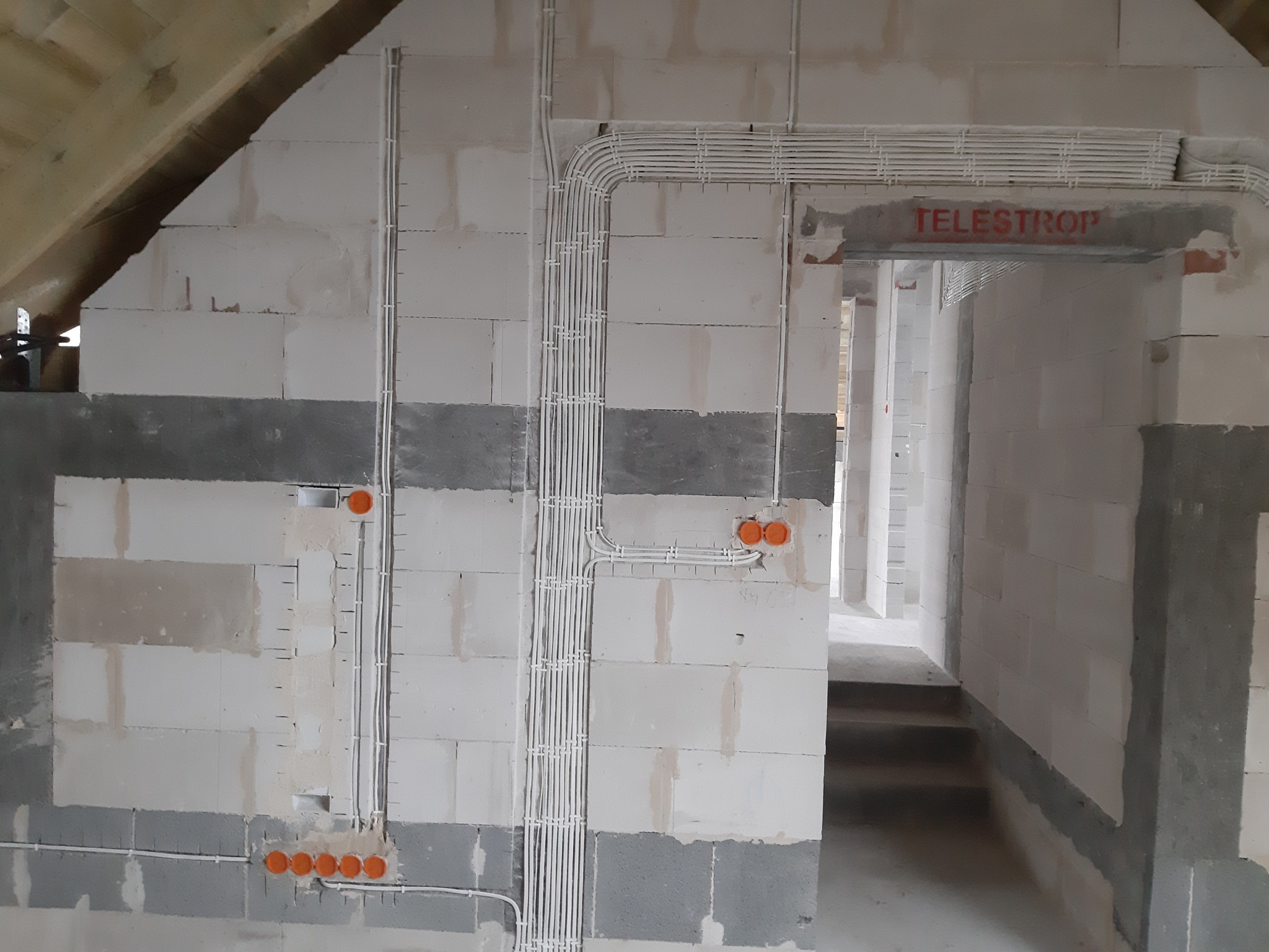 Surowe wnętrze w trakcie budowy z widocznymi, starannie ułożonymi przewodami elektrycznymi w białych rurkach, prowadzącymi do pomarańczowych puszek instalacyjnych na ścianach z betonu komórkowego...