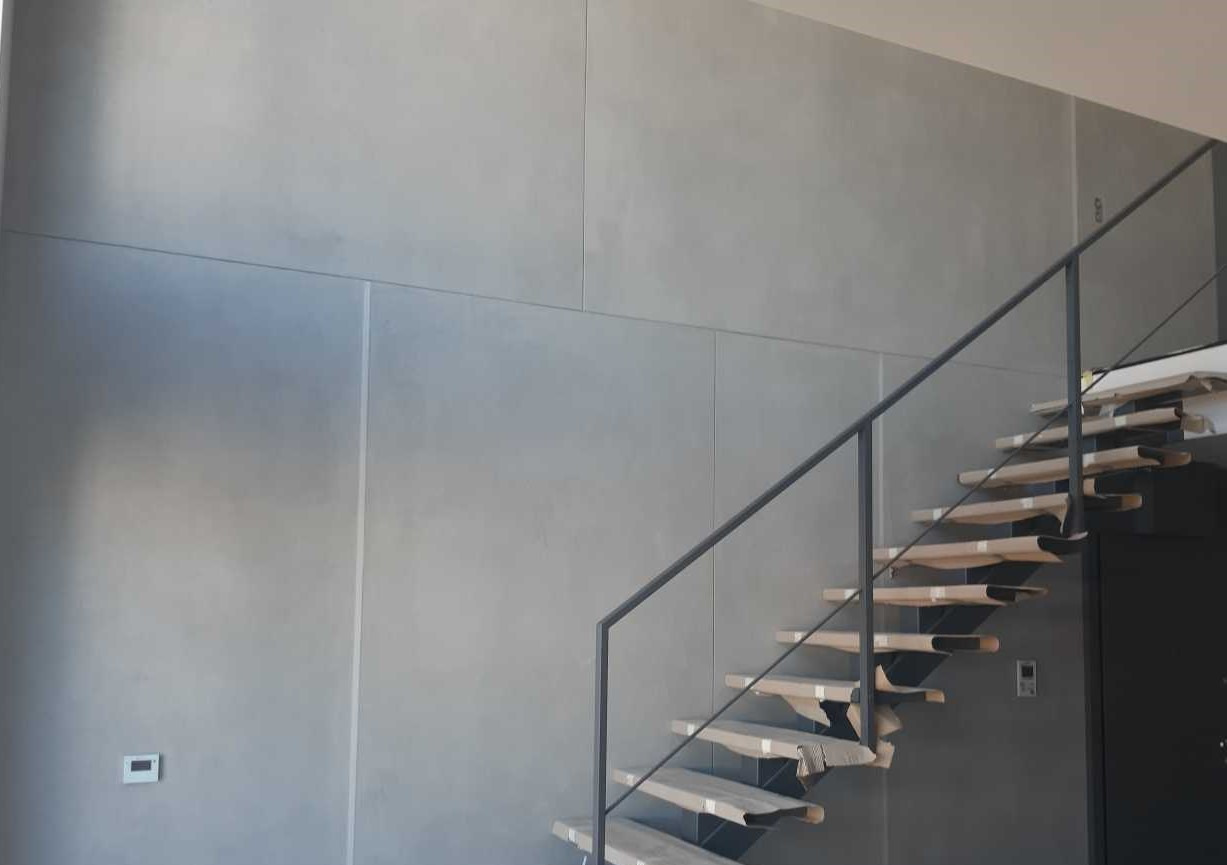Ściana wykończona szarym betonem architektonicznym z widocznymi podziałami na płyty, schody drewniane zabezpieczone kartonem i czarna metalowa balustrada.