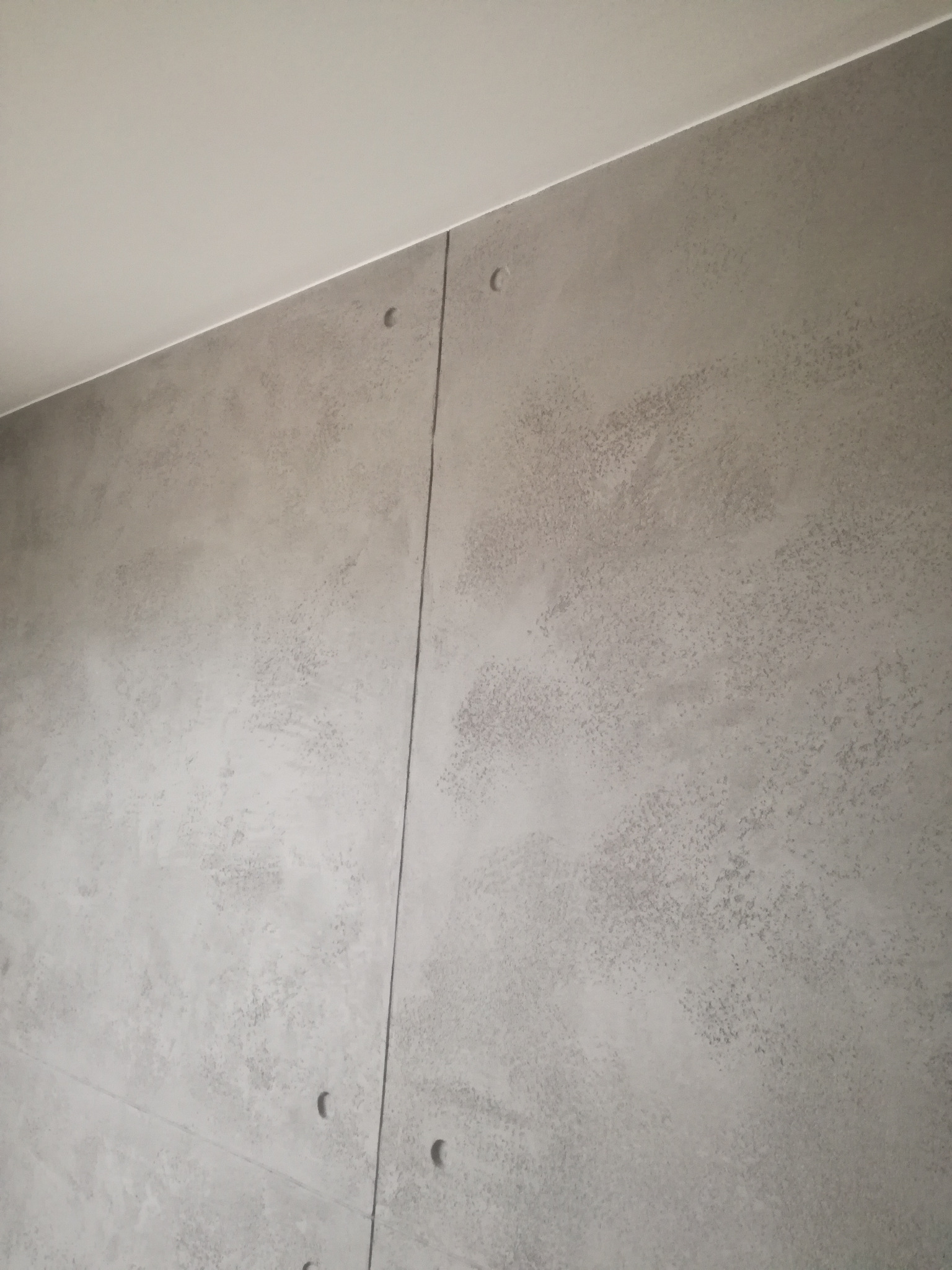 Ściana z szarego betonu architektonicznego z widocznymi łączeniami płyt i śladami po szpachlowaniu, oświetlona od góry.