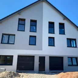 Dwukondygnacyjny dom jednorodzinny z dwiema bramami garażowymi, wykończony w odcieniach szarości i bieli, z czarnymi ramami okiennymi i drzwiami wejściowymi, widoczny stos materiałów budowlanych...