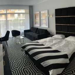 Nowoczesny pok&oacute;j hotelowy z ł&oacute;żkiem z czarno-białą pościelą, czarną sofą i geometrycznym dywanem, oświetlony światłem dziennym wpadającym przez okno.