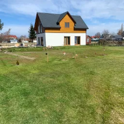 Nowoczesny dom jednorodzinny z białą elewacją i drewnianą górną częścią, ciemnoszary dach, widok z trawnika, w tle inne domy i błękitne niebo z chmurami.