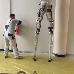 Dwóch mężczyzn w kombinezonach roboczych szpachluje ścianę w pomieszczeniu, jeden na szczudłach, drugi na podłodze, widoczne narzędzia i wiaderka na podłodze zabezpieczonej folią.