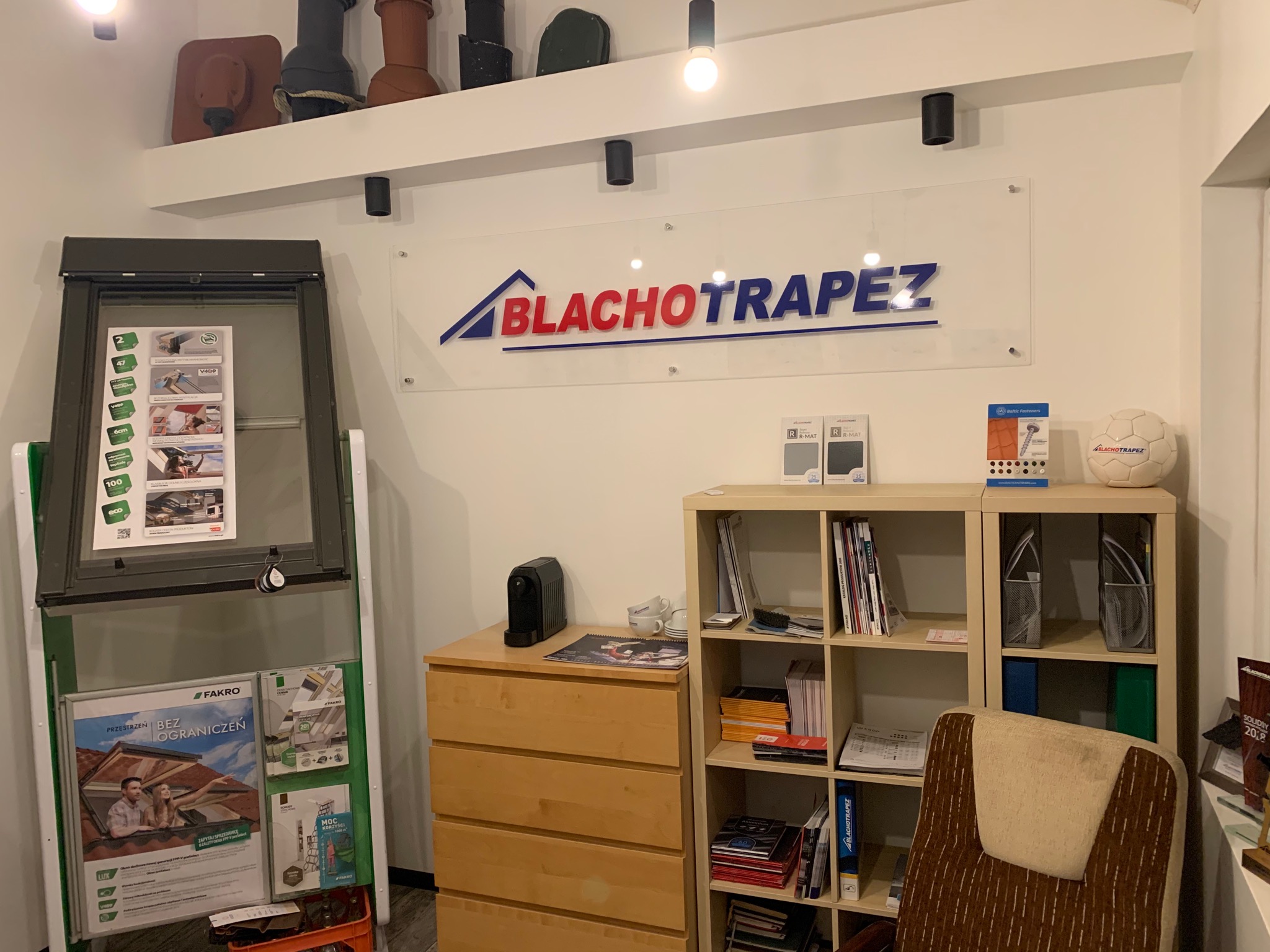 Wnętrze biura z ekspozycją okna dachowego Fakro na stojaku, katalogami i próbkami materiałów, nad biurkiem szyld firmy Blachotrapez i ekspozycja z modelami dachówek na półce.
