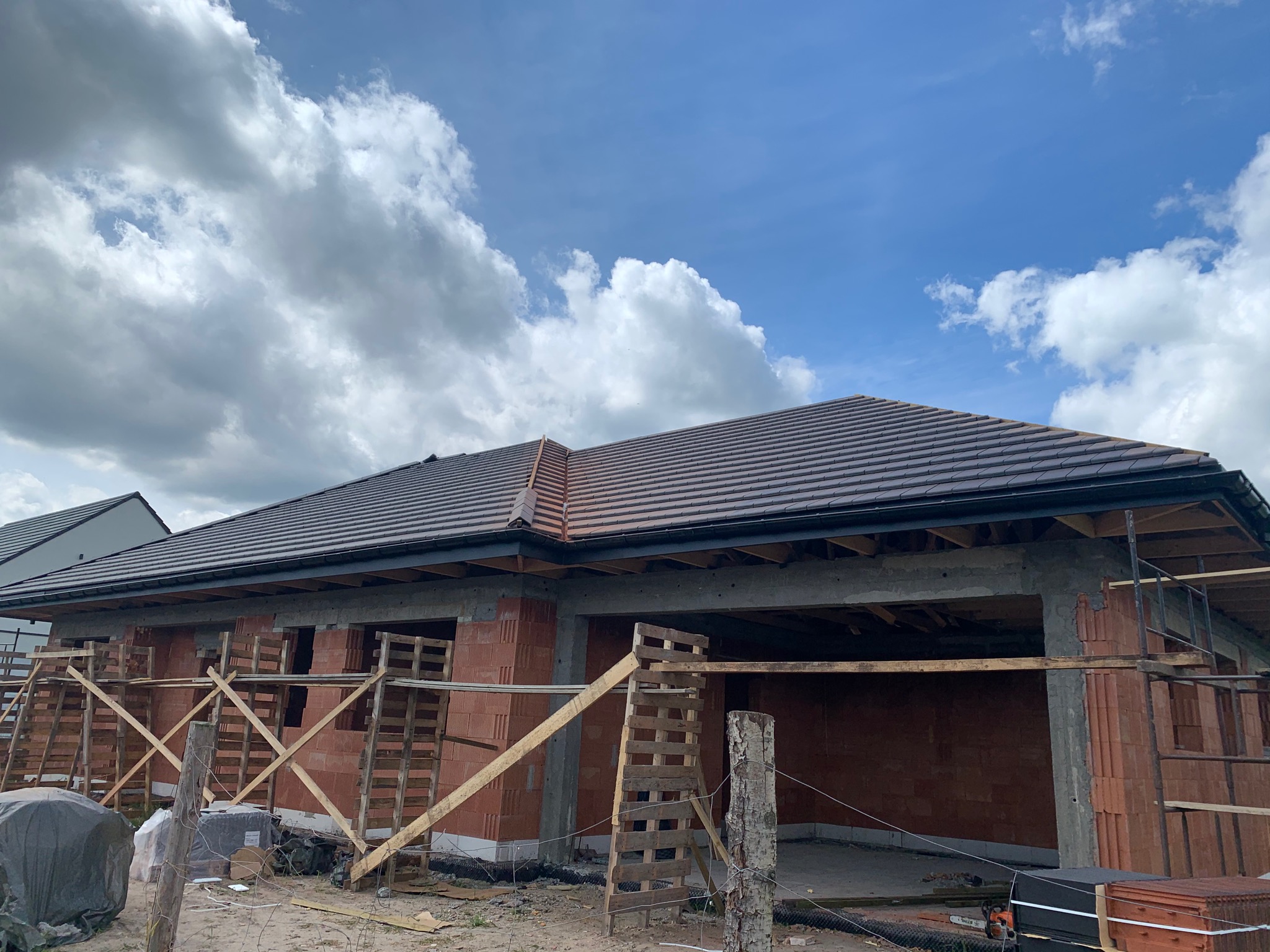 Dom w trakcie budowy, z ukończonym dachem z brązowej dachówki ceramicznej, widoczne rusztowania z drewnianych palet, ściany z pustaków ceramicznych, błękitne niebo z chmurami w tle.