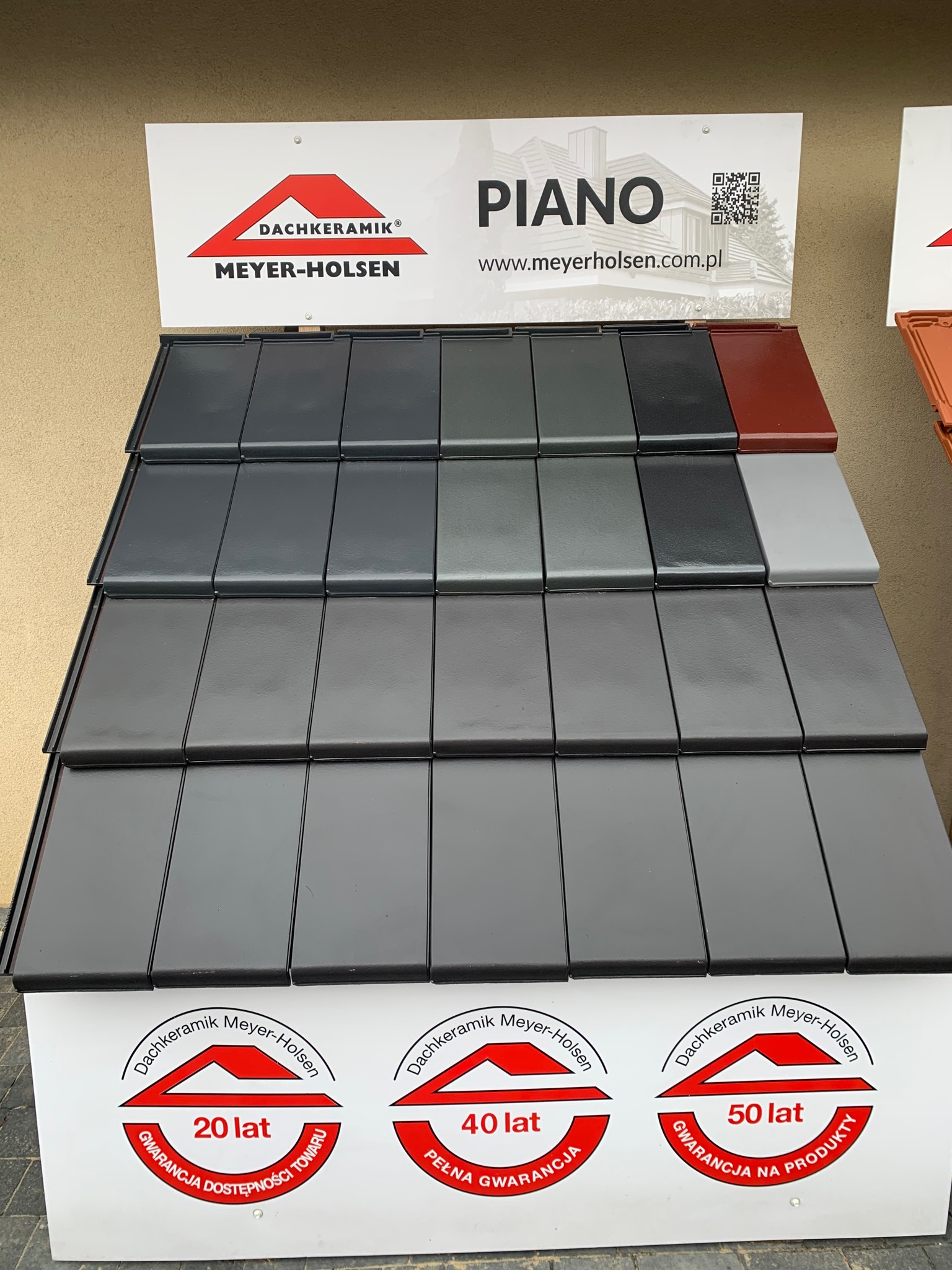 Ekspozycja różnych kolorów i wzorów ceramicznych elementów na dach, marki Meyer-Holsen, z informacją o długoletniej gwarancji.