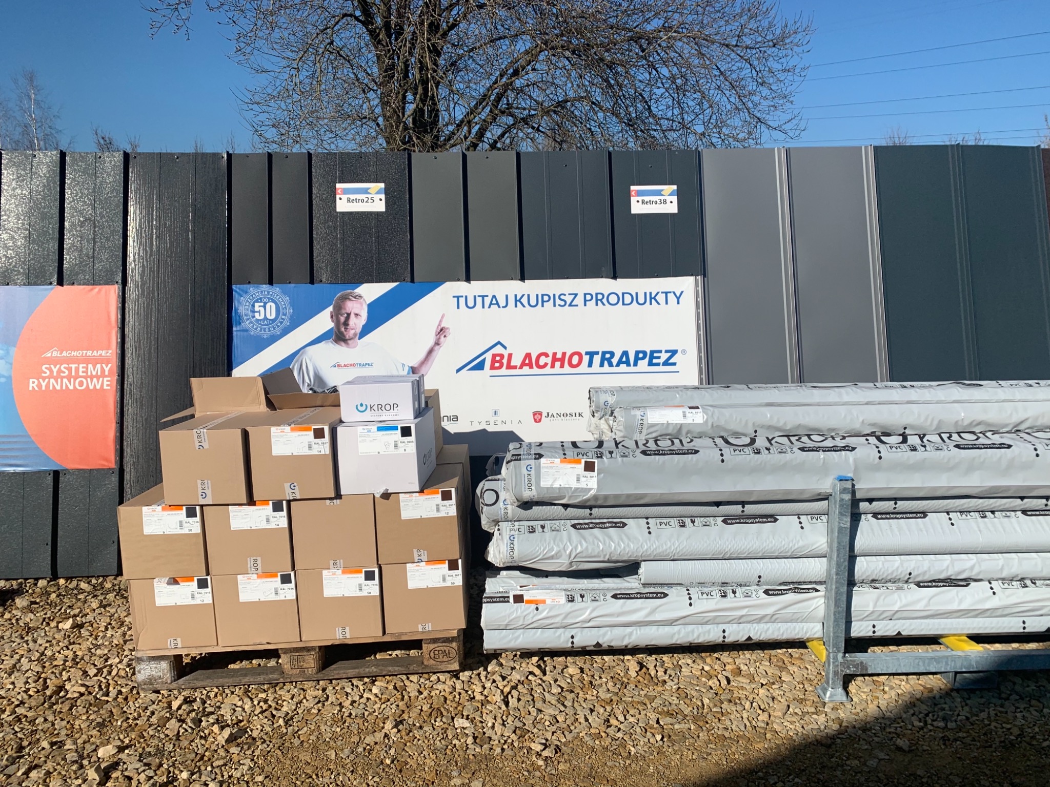 Stosy zapakowanych arkuszy i kartonów z produktami budowlanymi na palecie przed ekspozycją próbek różnych kolorów i faktur, firmowane logiem Blachotrapez oraz Krop.