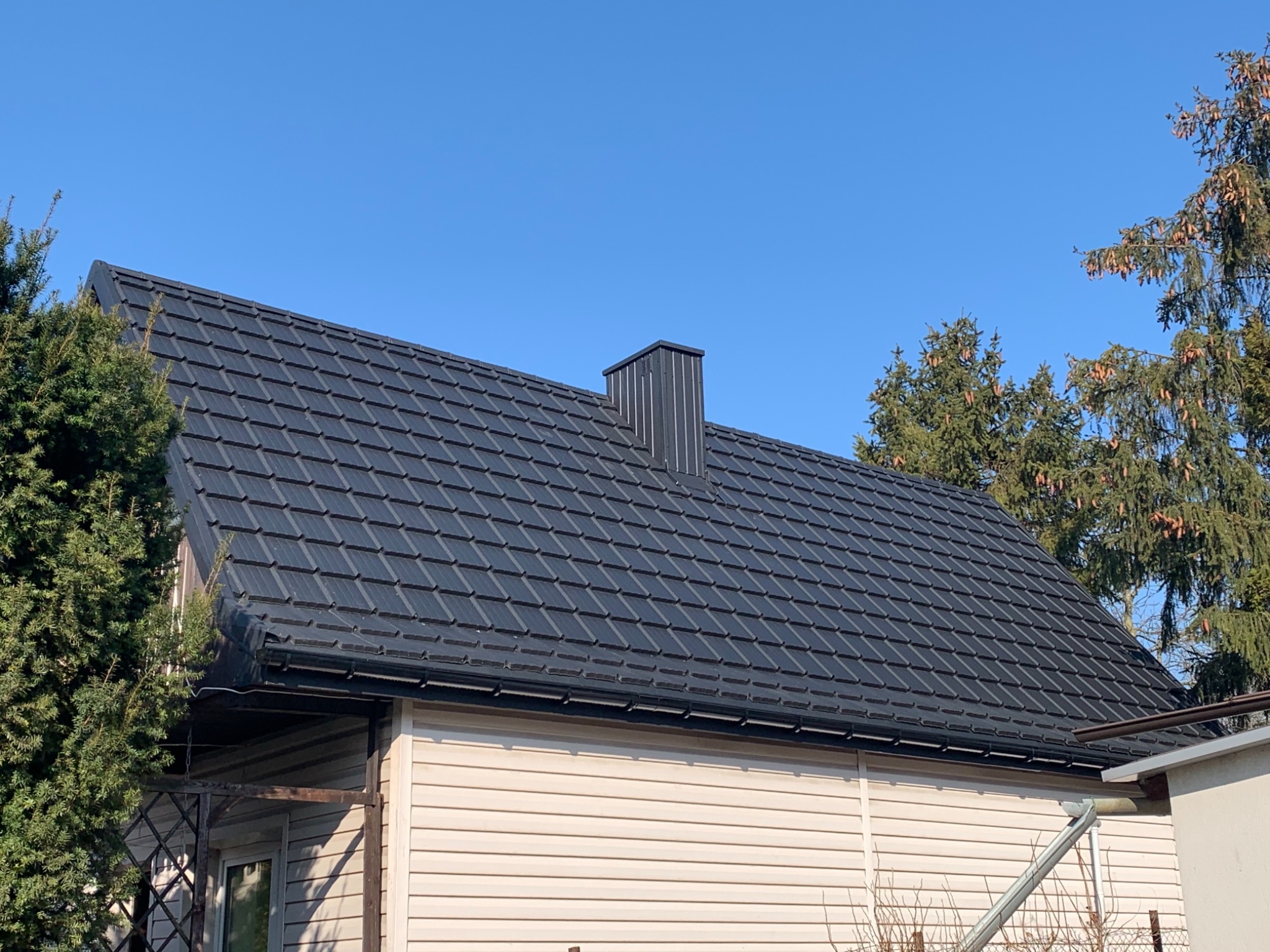 Czarny, metalowy dach z kominem na jasnym domu z sidingiem, na tle błękitnego nieba i zielonych drzew.