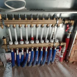 Rozdzielacz ogrzewania podłogowego z rurami w izolacji, zamontowany w metalowej szafce, widoczne przyłącza i zawory, ściana z surowej cegły w tle.