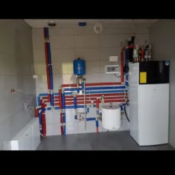 Kotłownia z piecem, zasobnikiem ciepłej wody i rozbudowaną instalacją z rur w kolorach czerwonym i niebieskim, na tle białych płytek.