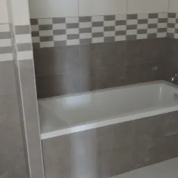 Wykończona łazienka z białą wanną, toaletą i szaro-białymi płytkami w szachownicę na ścianach, widoczny fragment instalacji wodnej.