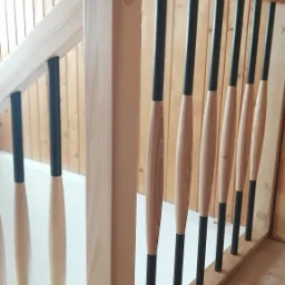 Jasne drewno i czarne elementy nowoczesnej balustrady schodowej z ozdobnymi wrzecionami, widok z dołu, drewniana boazeria w tle.