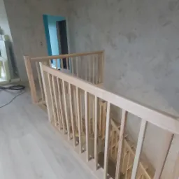 Jasne, surowe drewno balustrady schodowej w trakcie montażu, widok z góry. W tle ściany w odcieniach szarości i błękitne drzwi. Podłoga w jasnym odcieniu drewna.