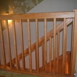 Z.P.U Marek Talaśka - Jasna, drewniana balustrada schodowa z pionowymi szczeblami w domu w Osie. Widoczne schody i fragment ściany z wzorzystą tapetą. Ciepłe oświetlenie.