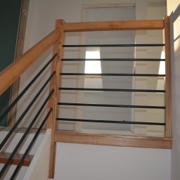 Z.P.U Marek Talaśka - Jasne drewno i czarne pręty tworzą nowoczesną balustradę schodową. Widoczne fragmenty schodów i ściany w trakcie wykończenia. Ujęcie z dołu, podkreślające geometrię.