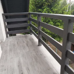 Szary balkon z drewnopodobnymi płytkami i nowoczesną, poziomą balustradą na tle zieleni.