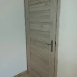 Zamknięte, wewnętrzne drzwi z drewnianą okleiną w odcieniach szarości, z klamką w kolorze srebrnym, zamontowane w Osie.