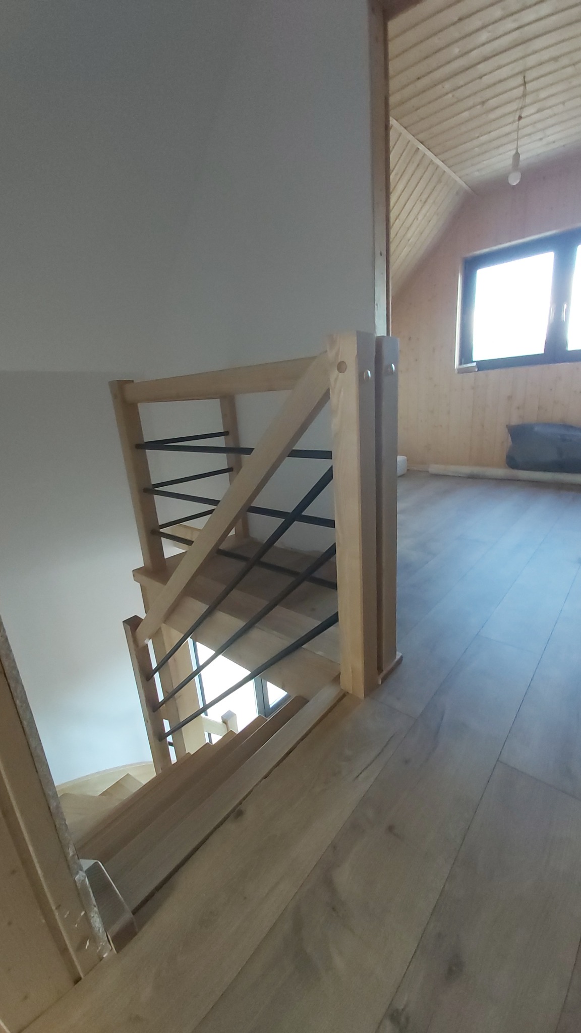 Nowoczesne schody z jasnego drewna z czarnymi, metalowymi prętami balustrady, widok z góry na dół, z drewnianym wykończeniem poddasza w tle.