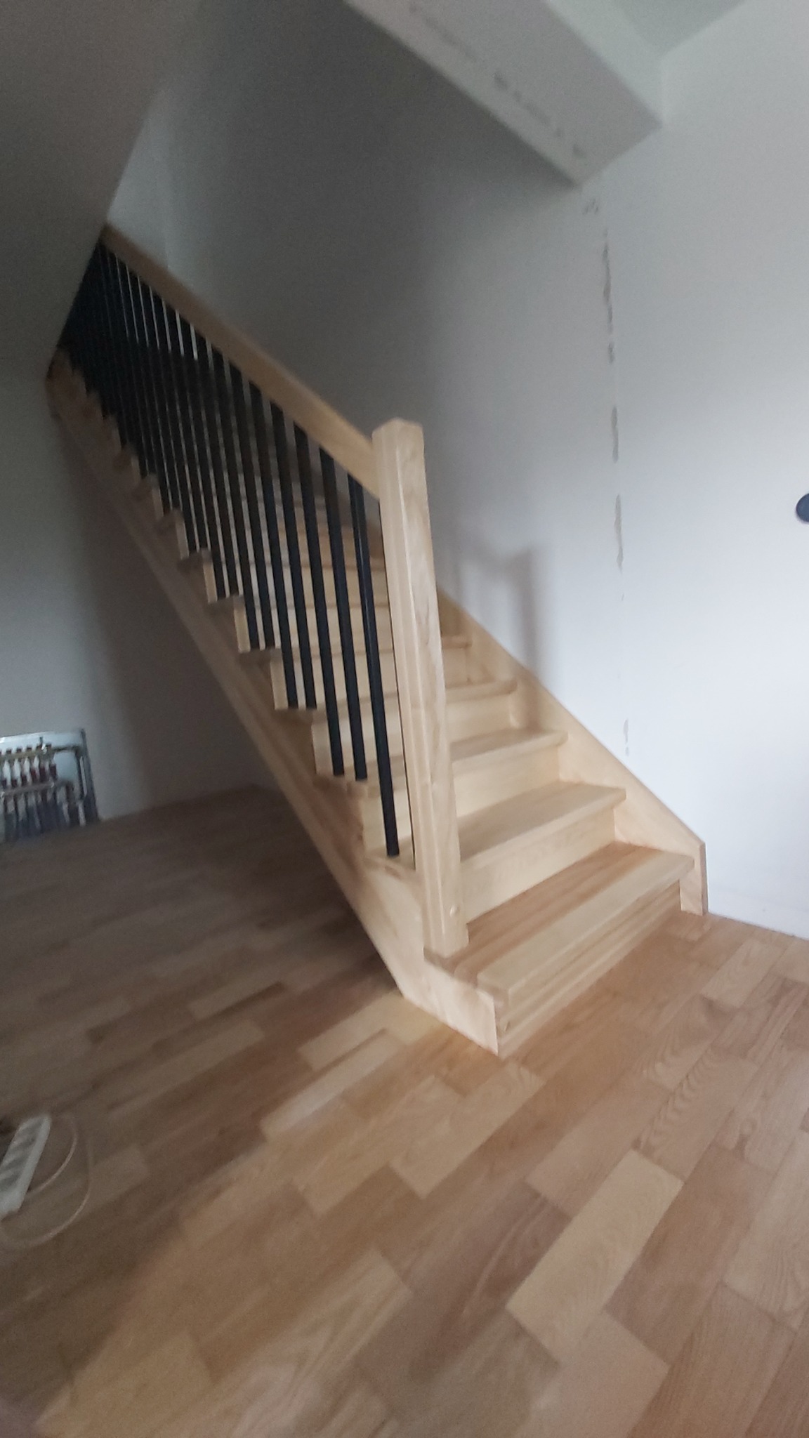 Jasne schody z drewnianymi stopniami i czarnymi, metalowymi balustradami, w jasnym wnętrzu. Widoczne drewniane podłogi i fragment ściany.