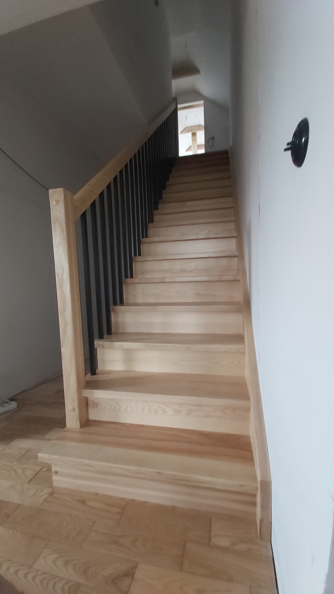 Jasne, drewniane schody z czarną, metalową balustradą w nowoczesnym wnętrzu. Widok z dołu schodów, z widocznym fragmentem podłogi i ściany.