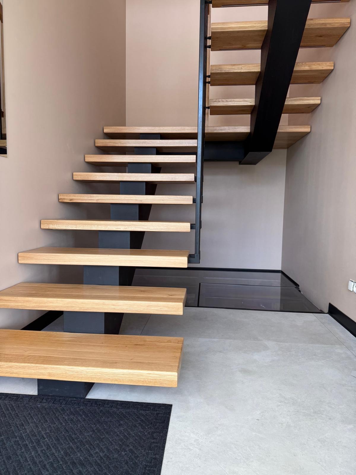 Nowoczesne schody drewniane z metalową konstrukcją nośną i ukrytą piwniczką pod schodami, wykończone w minimalistycznym stylu, z dywanikiem na podłodze.