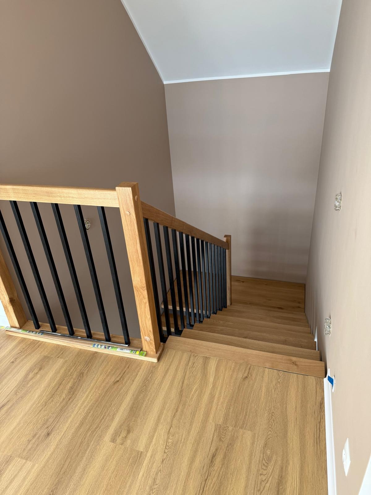 Drewniane schody z czarną balustradą w nowoczesnym wnętrzu. Jasne drewno schodów i podłogi kontrastuje z ciemnymi elementami balustrady. Widok z góry.