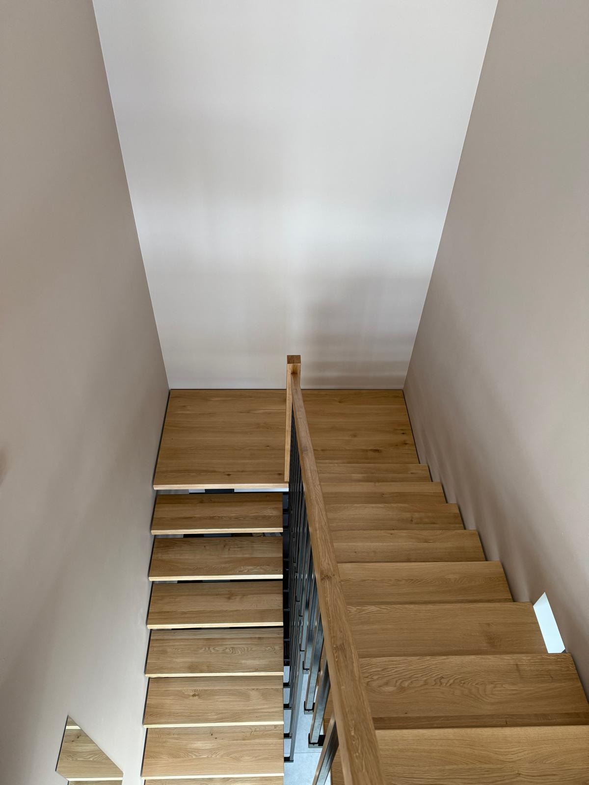 Nowoczesne schody z jasnego drewna, widok z góry. Minimalistyczna balustrada z ciemnego metalu. Schody prowadzą w górę, wzdłuż ściany o neutralnym odcieniu.