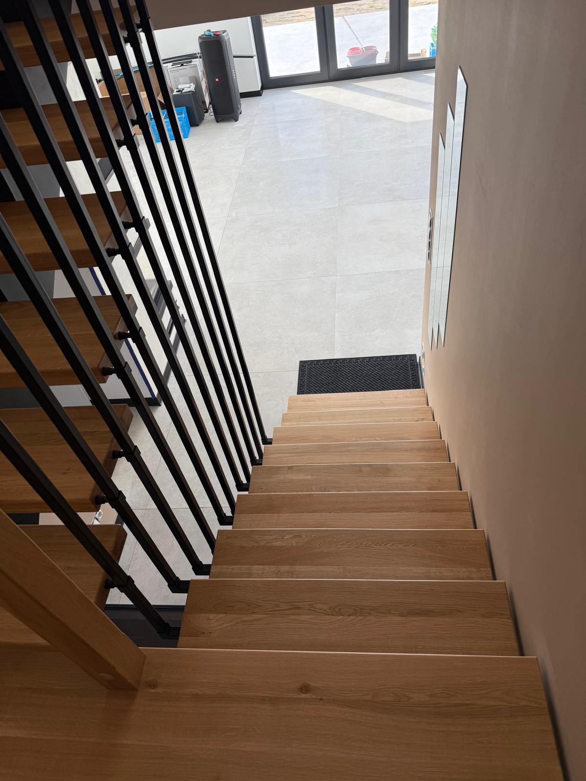 Nowoczesne schody z drewnianymi stopniami i czarną, metalową balustradą. Widok z góry na jasne wnętrze z płytkami i wejściem.